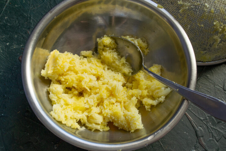 Cooked potatoes being passed through a potato ricer into a white bowl to create a fine texture for gnocchi.