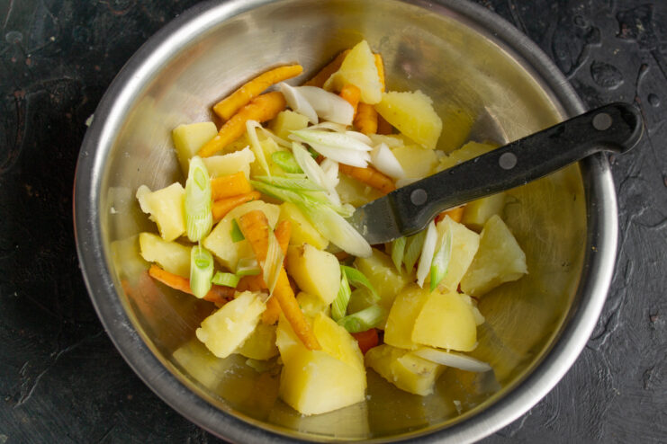 A bowl with boiled potatoes and carrots, with chopped green onions being added.