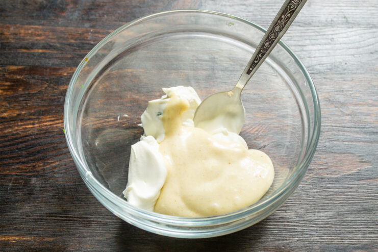 Adding mayonnaise to the salad dressing base Adding a dollop of mayonnaise to the cream cheese and sour cream mixture in a bowl.