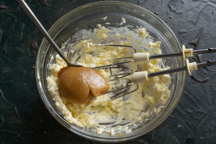 Gradually adding dulce de leche to the whipped butter. A spoon adding a portion of dulce de leche to the whipped butter in a bowl.