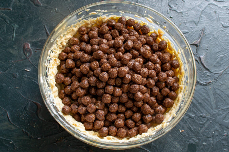 Adding the main crunchy ingredient: chocolate cereal balls. A hand pouring chocolate cereal balls from a box into a large bowl.
