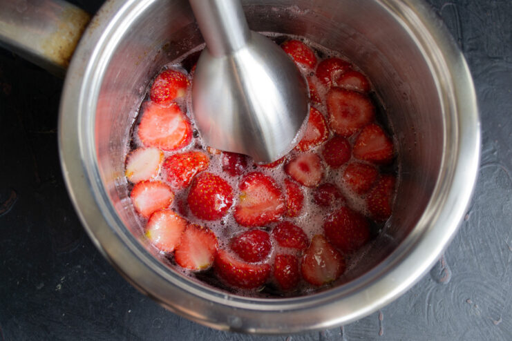 Step 7: Blending cooked strawberries into a smooth puree. Using an immersion blender to puree the cooked strawberries in a saucepan.
