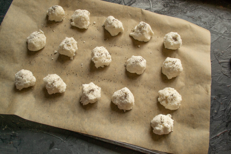 Sprinkling the Cookies with Poppy Seeds Before Baking Raw coconut cookie dough balls on a baking sheet being sprinkled with poppy seeds.