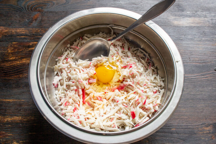 Binding the Mixture: Adding an Egg to Grated Crab Sticks - Essential for holding the cutlets together. A bowl with grated crab sticks and a raw egg being added to it.