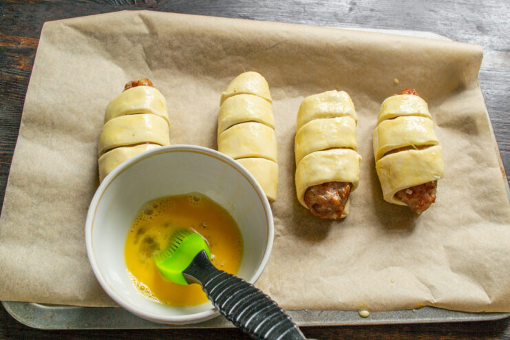 A hand using a silicone brush to apply egg wash to the pastry-wrapped patties.