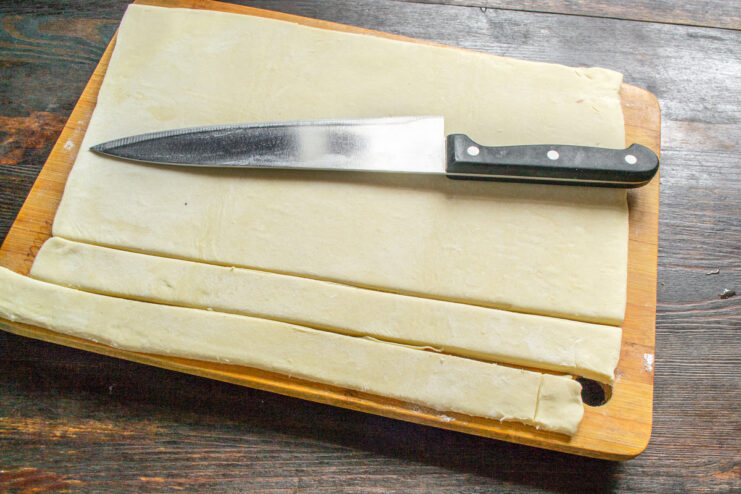 A sheet of puff pastry being cut into long, even strips with a knife.