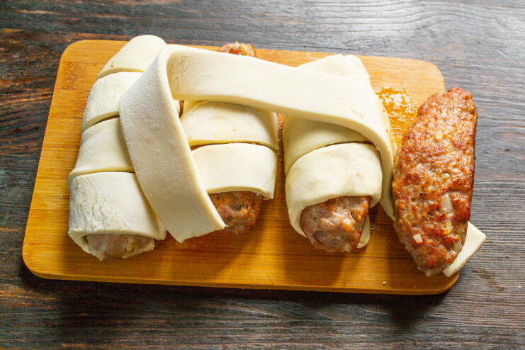 A seared meat patty being carefully wrapped with a strip of puff pastry.
