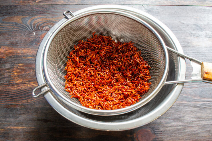Step-by-step: How to properly rinse soaked red rice before cooking. Soaked red rice being rinsed in a metal colander under running water.