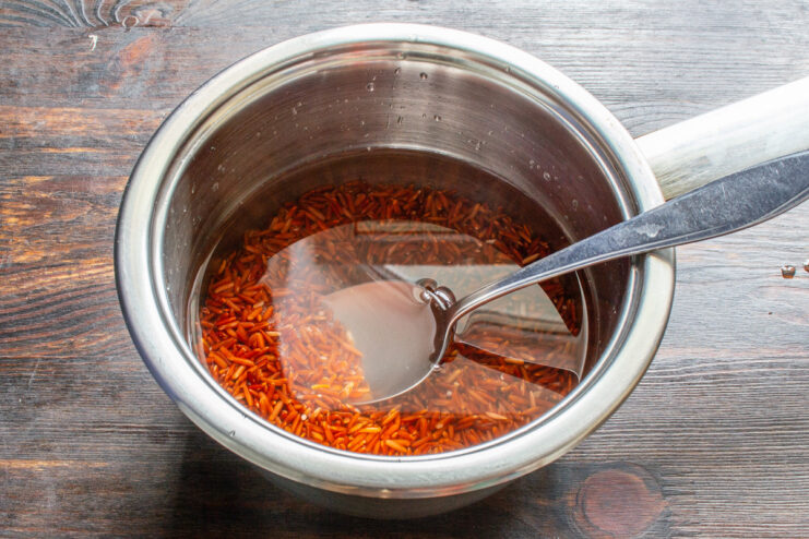 Adding the correct amount of water to cook perfect red rice. A hand pouring water from a measuring cup into a saucepan with red rice.
