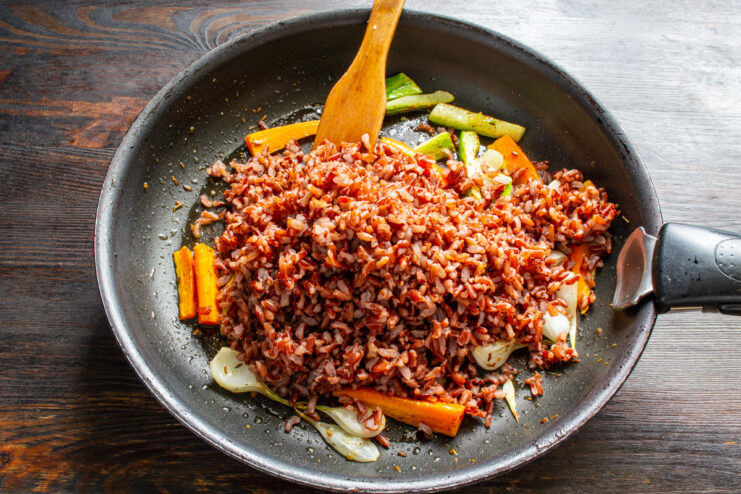 Combining cooked red rice with sautéed vegetables. Cooked red rice being added to a pan of sautéed vegetables.