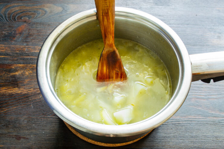 Adding hot broth to the sautéed onions and potatoes in a saucepan. Hot broth being poured into a saucepan with sautéed onions and potatoes.