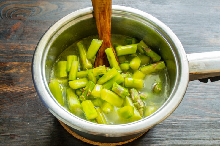 Adding chopped asparagus to the simmering soup base. Chopped green asparagus being added to the saucepan with potatoes and broth.