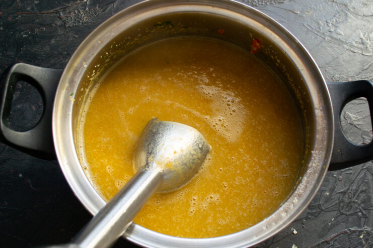 An immersion blender being used to puree the cooked vegetable soup in a pot.
