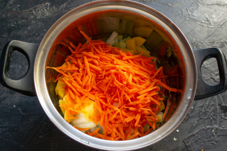 Grated carrots being added to a pot with potatoes, onions, and celery.