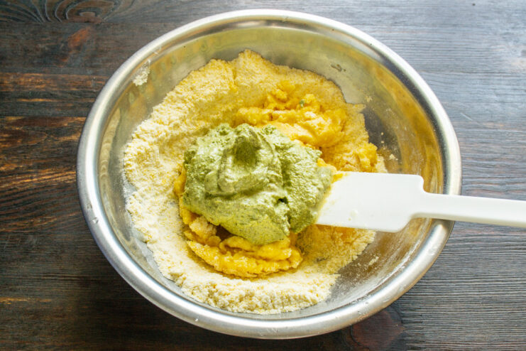 Adding a Flavorful Pesto Boost to the Fritters A spoonful of green pesto sauce being added to the corn fritter batter.
