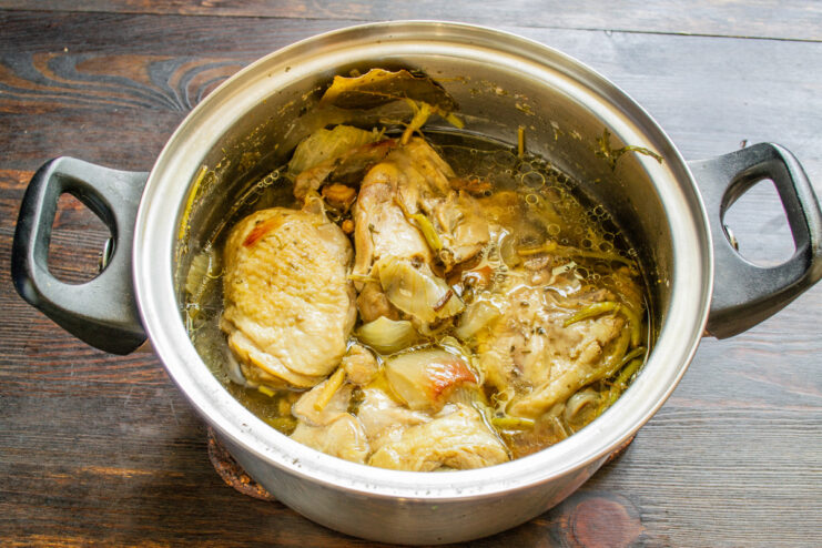 Chicken thighs simmering in a pot with aromatics like onion, garlic, and herbs.