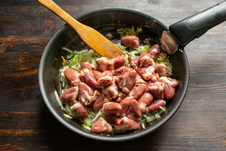Adding the prepared chicken hearts to the sautéed aromatics. Prepared chicken hearts being added to a skillet with sautéed onions.