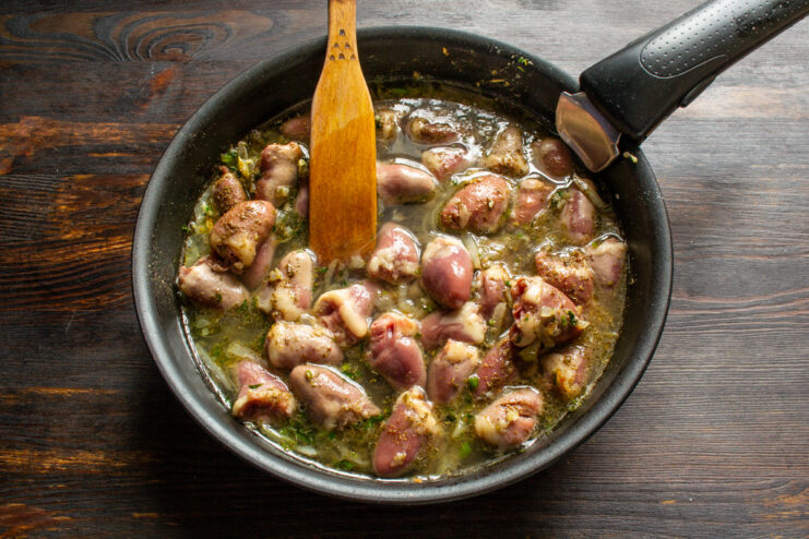 Searing the chicken hearts with spices before adding liquid for stewing. Pouring chicken broth into a skillet with chicken hearts and spices.