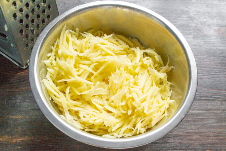 Step 1: Grating Potatoes for the Kolduny Mixture A hand grating a peeled potato into a glass bowl using a box grater.