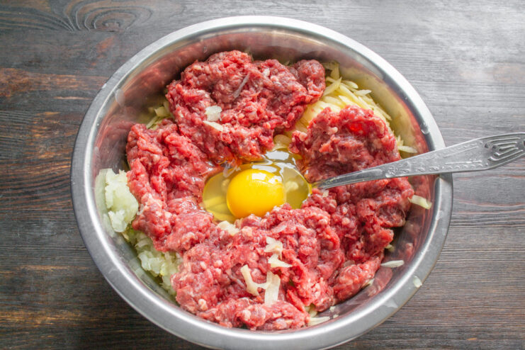 Binding the Ingredients: Adding an Egg to the Mixture A large chicken egg cracked into the bowl with grated potatoes, onion, and ground beef.