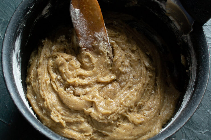 Creating a choux-style dough by mixing flour into the hot honey syrup. Mixing wheat flour into the hot syrup in a saucepan to form the dough.