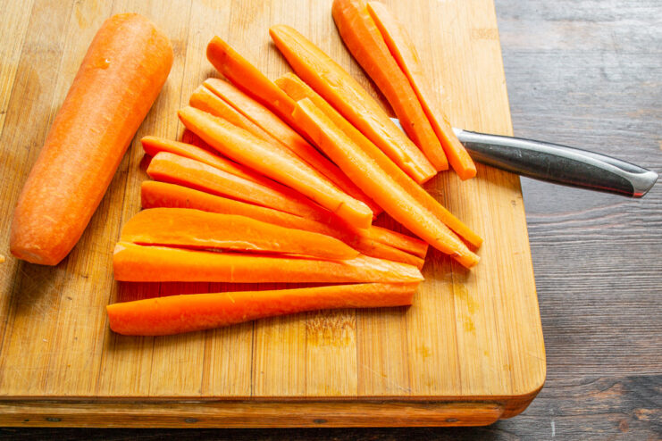 Peeled carrots being cut into long, thick sticks on a cutting board.