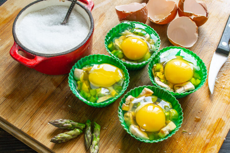 Adding Fresh Eggs to the Vegetable Base A large, bright-yolked egg cracked into a mold over asparagus and mushrooms.