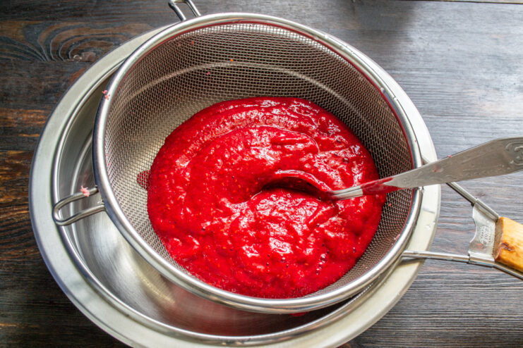 Step 2: Straining raspberry puree to remove seeds for a smoother texture. A hand using a spoon to press raspberry puree through a fine-mesh sieve over a bowl.