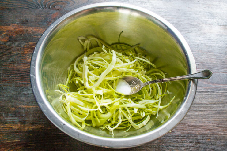 Shredded white cabbage in a glass bowl being sprinkled with salt.