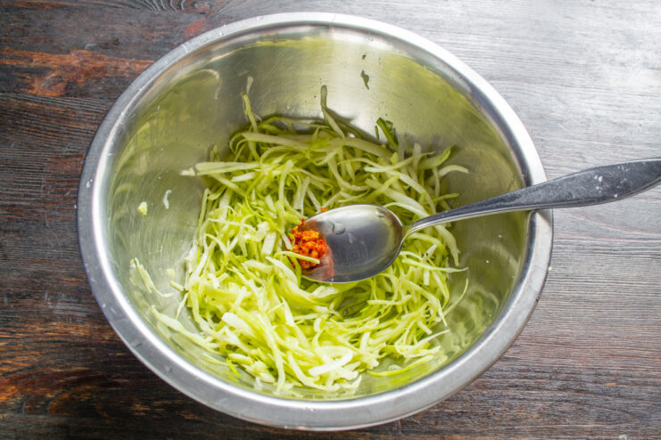 A hand mixing raw adjika paste into salted cabbage in a glass bowl.