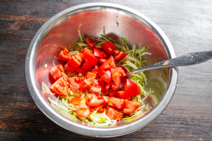 Diced red tomatoes being added to the cabbage mixture in a glass bowl.