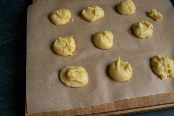 Portioning the Choux Dough Before Frying Spoonfuls of raw choux pastry dough portioned out onto a sheet of parchment paper.