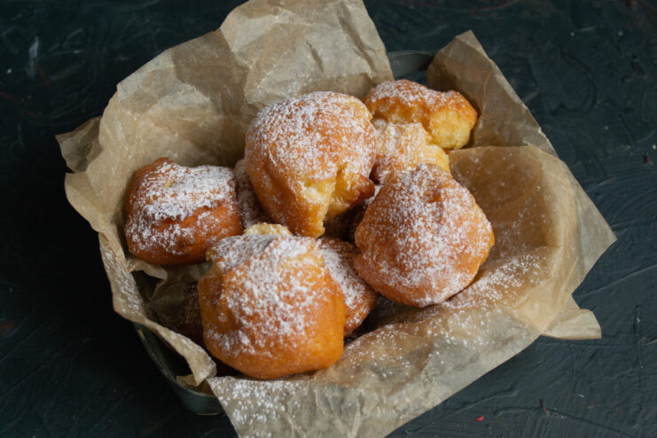 Serving Freshly Fried Choux Pastry Doughnuts A close-up of golden-brown choux pastry doughnuts on a plate, being dusted with powdered sugar.