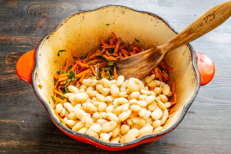 Combining Cooked Beans with Sautéed Vegetables Cooked white beans being added to a pot with sautéed carrots.
