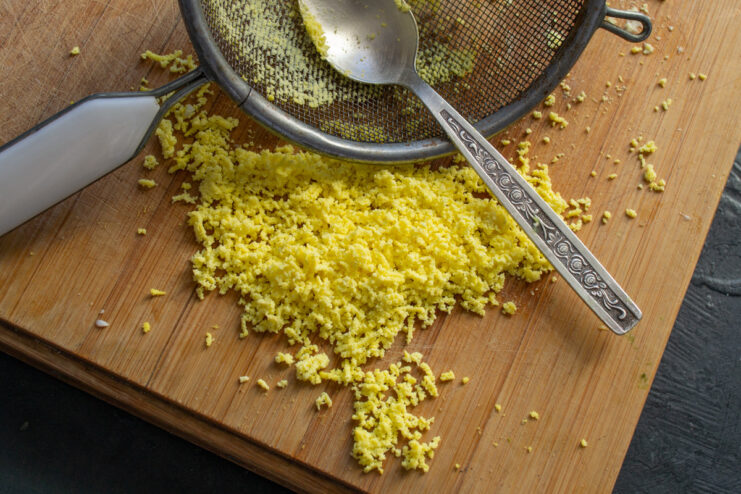 Preparing Grated Egg Yolk for Garnish A hand pressing a hard-boiled egg yolk through a small metal sieve over a bowl.