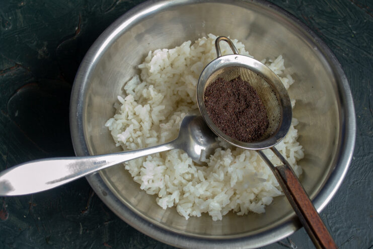 A bowl of cooked rice with poppy seeds and seasoning being added.