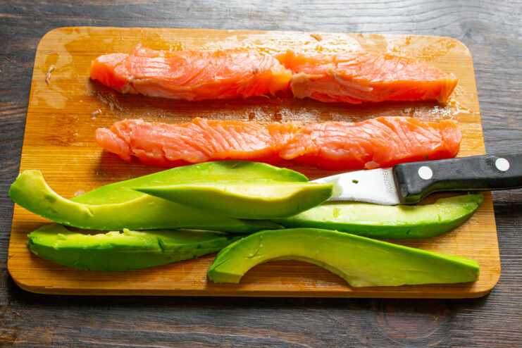 How to Slice Avocado for Sushi A hand slicing a peeled avocado half into long, thin strips on a cutting board.