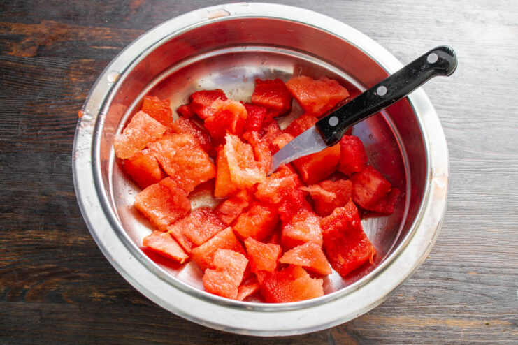 Pro tip: Cut watermelon over the bowl to save all the juice for the dressing. Hands chopping fresh watermelon cubes over a white mixing bowl.