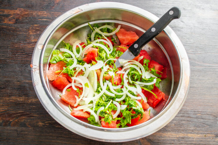 Thinly sliced onions provide a sharp contrast to the sweet watermelon. A hand slicing a yellow onion into thin half-rings on a cutting board.