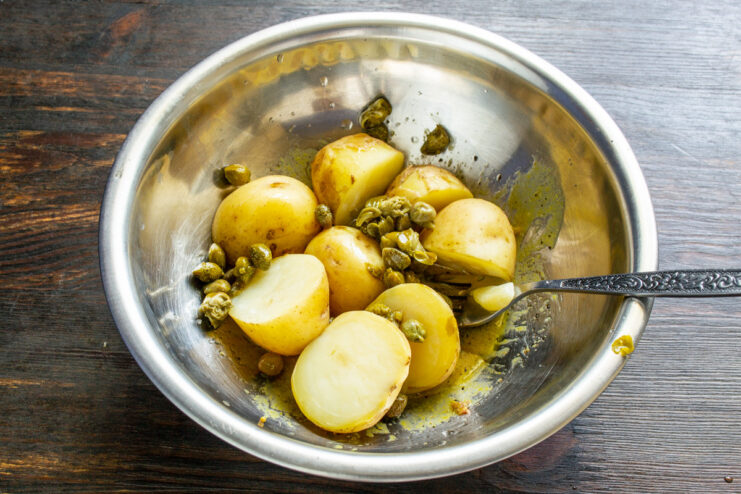 Adding briny capers for a burst of tangy flavor. Adding a spoonful of capers to the potato salad in a bowl.