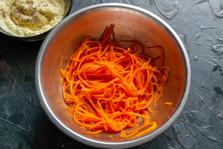 Dressing the Korean-style carrots with olive oil to keep them fresh. A bowl of bright orange Korean-style carrots being drizzled with olive oil.