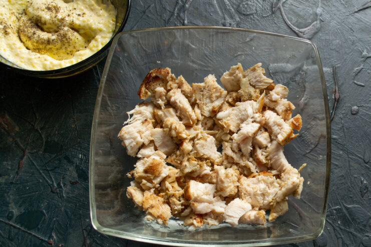 Dicing the cooked chicken for the first layer of the salad. Cooked and cooled chicken breast being cut into small cubes on a cutting board.