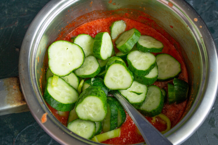 Sliced cucumbers and hot peppers being added to the simmering tomato sauce in a pot.