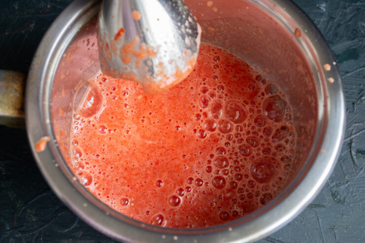 An immersion blender pureeing a mixture of tomatoes, peppers, and garlic in a pot.