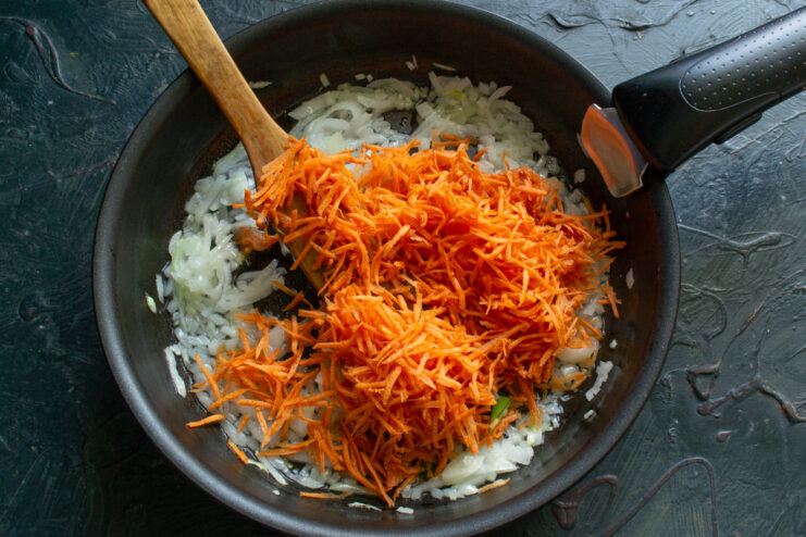 Adding grated carrots to the onions to build the soup's flavor Grated carrots being added to the sautéed onions in a pan.