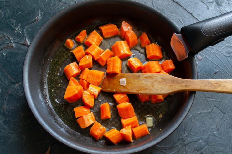Sautéing Carrots to Build Flavor Diced carrots being sautéed in a black skillet with a wooden spatula.