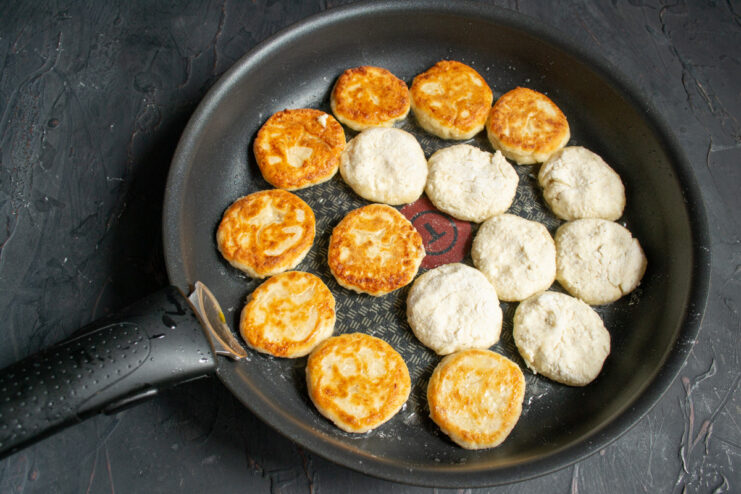 Flipping Syrniki to Cook Until Golden Brown Syrniki being fried in a pan, showing a golden-brown crust on one side.