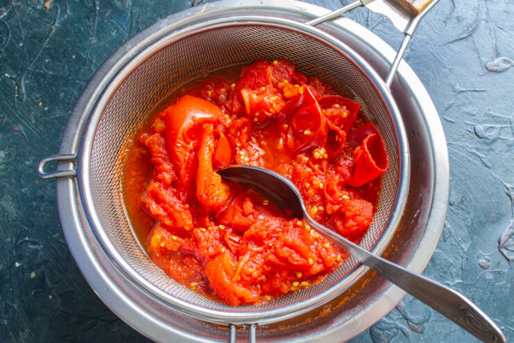 Sieving the Tomatoes: How to Get a Silky Smooth Sauce Cooked tomatoes being pressed through a large sieve with a spoon to separate the pulp from skins and seeds.