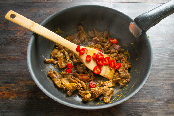 Sliced red chili peppers being added to the seared beef in a pan.