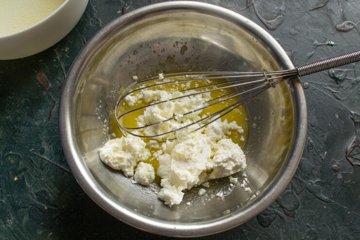 Adding Farmer's Cheese (Tvorog) to the Mixture Adding a portion of crumbly tvorog (farmer's cheese) to the mixing bowl.